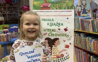 Uśmiechnięta dziewczynka w blond włosach stoi w bibliotece w Zblewie trzymając sw&oacute;j dyplom, za nią stoi tablica z plakatami dotyczącymi projektu Mała Książka Wielki Człowiek