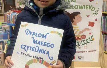 Chłopiec w ciemnej karnacji w granatowej kurtce stoi w bibliotece w Zblewie pokazując sw&oacute;j dyplom. Za nim stoi tablica z plakatami dotyczącymi projektu Mała Książka Wielki Człowiek