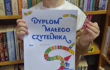 Uśmiechnięty chłopiec w ciemnych włosach stoi w bibliotece w Borzechowie trzymając dyplom i kartę Małego Czytelnika.