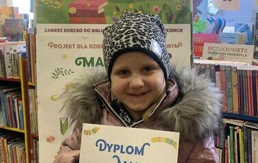 Uśmiechnięta dziewczynka w r&oacute;zowej kurtce stoi trzymając sw&oacute;j dyplom, za nią stoi tablica korkowa z plakatami dotyczącymi projektu Mała Książka Wielki Człowiek