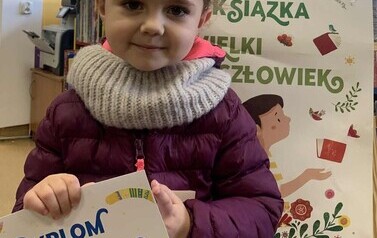 Dziewczynka w kr&oacute;ciutkich ciemnych włosach w fioletowej kurtce stoi trzymając dyplom, za nią stoi tablica z plakatem dotyczącym projektu Mała Książka Wielki Człowiek