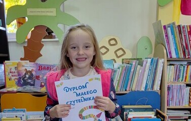 Dziewczynka w blond włosach do ramion siedzi na krzesełku w bibliotece w Pinczynie i pokazuje sw&oacute;j dyplom. Uśmiecha się.