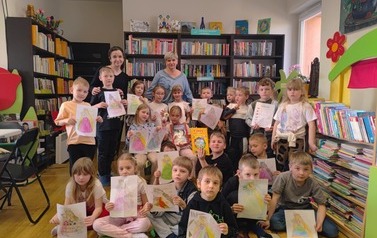 Zdjęcie grupowe dzieci ze swoją Panią i z Panią bibliotekarką.  Dzieci trzymają swoje pokolorowane prace plastyczne pokazując je.
