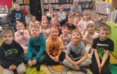 Dzieci siedzą na dywanie, za nimi na krzesełku siedzi ich Pani.