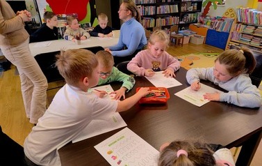 Dzieci siedzą przy dw&oacute;ch stołach i wykonują prace plastyczne o wiośnie. Przy jednym stole siedzi ich Pani, a obok białego stołu stoi nauczycielka