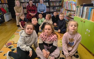 Dzieci siedzą na dywanie, a za nimi na małych trzech krzesełkach siedzą ich Panie.