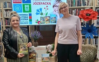 Przy stoliku stoją pisarka i kierowniczka biblioteki, kt&oacute;ra trzyma zakupioną książkę. Za nimi wyświetla się na ekranie plakat ze spotkania i stoi  stojak ze sztucznymi kwiatami.
