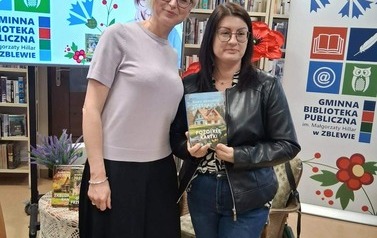 Pisarka stoi z czytelniczką, kt&oacute;ra trzyma zakupioną ksiązkę. Za nimi stoi duży ekran z wyświetlanym plakatem ze spotkania, stojak z sztucznymi kwiatami i rolap z logiem biblioteki.