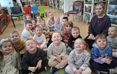 Dzieci siedzą na dywanie i się uśmiechają, obok nich siedzi ich Pani z Przedszkola