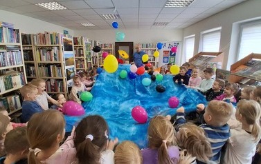 Dzieci stoją w dużym k&oacute;łku mając szeroko rozłożony duży materiał kt&oacute;rym podrzucają kolorowe balony.