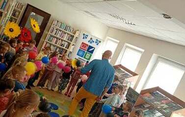 Dzieci stoją w k&oacute;łku trzymając kolorowe balony. W środku koła chodzi Pan Poeta.