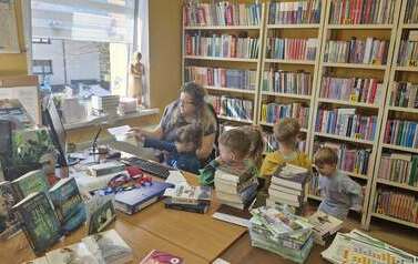 Dzieci kolejno podchodzą do biurka i z pomocą bibliotekarki klikają kodem kreskowym w książce do czytnika wprowadzając tym samym książkę na konto czytelnika.