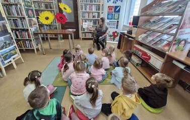 Dzieci siedzą na dywanie i siedziskach i patrzą w stronę bibliotekarki, kt&oacute;ra im czyta bajkę