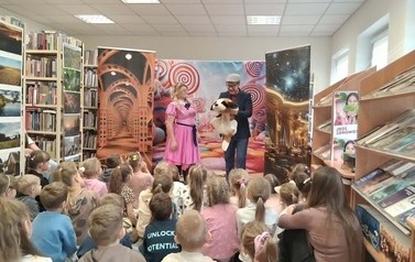 Dzieci siedzą na dywanie i patrzą na Zosię w r&oacute;żowej sukience i obok stojącego Feliksa trzymającego dużego pluszowego pieska.