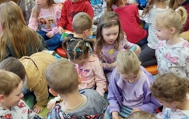 Duża grupa dzieci siedzi na dywanie i siedziskach i bawi się pluszowymi książeczkami.