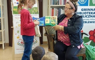Dzieci siedzą na siedziskach, z przodu bibliotekarka trzyma pluszową książeczkę "sklep" a obok dziewczynka trzyma siateczkę i bawi się w robienie zakup&oacute;w odklejając z książeczki poszczeg&oacute;lne elementy