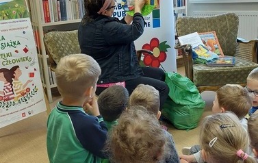 Dzieci siedzą na siedziskach i patrzą na bibliotekarkę, kt&oacute;ra rozkłada pluszową książeczkę.