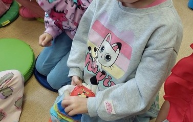 Dziewczynka w blond wlosach i w siwej bluzeczce bawi się pluszową książeczką, a obok niej siedzą inne dzieci.