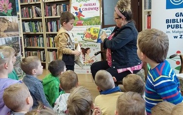 Dzieci siedzą na siedziskach, z przodu bibliotekarka trzyma pluszową książeczkę "sklep" a obok chłopiec trzyma siateczkę i bawi się w robienie zakup&oacute;w odklejając z książeczki poszczeg&oacute;lne elementy