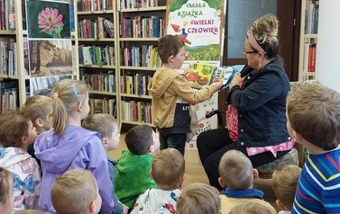 Dzieci siedzą na siedziskach, z przodu bibliotekarka trzyma pluszową książeczkę "sklep" a obok chłopiec trzyma siateczkę i bawi się w robienie zakup&oacute;w odklejając z książeczki poszczeg&oacute;lne elementy