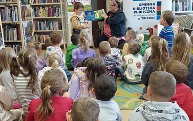 Dzieci siedzą na siedziskach, z przodu siedzi bibliotekarka trzymając pluszową książeczkę "sklep", a przy Niej stoi chłopiec trzymając małą siateczkę do zakup&oacute;w odklejając z książeczki poszczeg&oacute;lne elementy