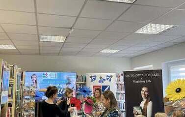 Kierowniczka biblioteki wręcza pisarce kwiaty i prezent. Dwie bibliotekarki z przodu robią zdjęcia. 