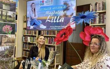 Przy stoliku okrągłym siedzi uśmiechnięta pisarka i kierowniczka biblioteki z teczką w dłoniach. Na stoliku są wyłożone pionowo książki pisarki. Wyżej nad głowami jest duży ekran z wyświetlanym plakatem ze spotkania.