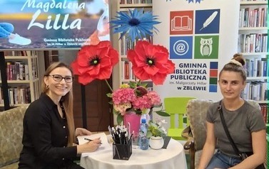 Pisarka przy stoliku podpisuje na książce dedykację czytelniczce, kt&oacute;ra siedzi obok. Obie się uśmiechają.