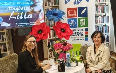Pisarka przy stoliku podpisuje na książce dedykację czytelniczce, kt&oacute;ra siedzi obok. Obie się uśmiechają.