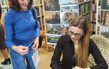 Pisarka przy stoliku podpisuje dedykację na książce i wstawia pieczątkę, obok stoi czytalniczka trzymająca torebkę papierową na książkę zakładkę.