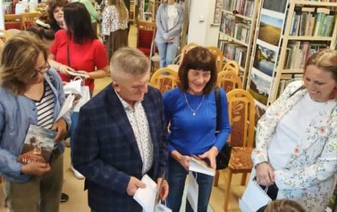 Przy stoliku pisarki, kt&oacute;ra podpisuje dedykacje na książkach stoją czytelnicy w kolejce z zakupionymi książkami. Reszta czytelnik&oacute;w rozmawia przy kawie i cieście.