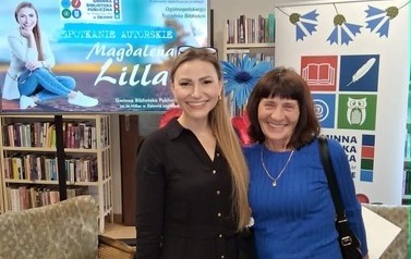 Pisarka stoi z czytelniczką, obie się uśmiechają. Za nimi stoi rolap z logiem biblioteki i ekran z wyświetlanym plakatem ze spotkania.