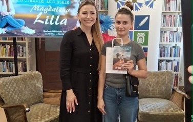 Pisarka stoi z czytelniczką, kt&oacute;ra trzyma zakupioną książkę. Za nimi rolap z logiem biblioteki i ekran z wyświetlanym plakatem ze spotkania.