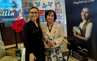 Pisarka stoi z czytelniczką , kt&oacute;ra trzyma zakupioną książkę. Po prawej stoi rolap z wizerunkiem pisarki i rolap z logiem biblioteki, a po lewej ekran z wyświetlanym plakatem ze spotkania.