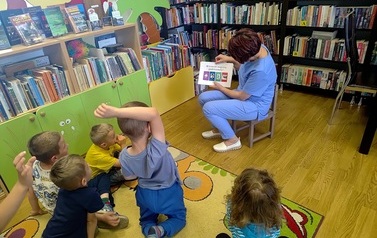 Pani Gabrysia z przodu pokazuje dzieciom kartkę z pytaniem do wersza "Lokomotywa", a dzieci siedzą na dywanie i niekt&oacute;re podnoszą rękę w celu udzielenia odpowiedzi