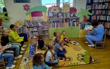 Dzieci siedzą na dywanie i krzesełkach i patrzą na Panią Gabrysię, ktora im czyta bajkę obrazkami skierowaną do dzieci