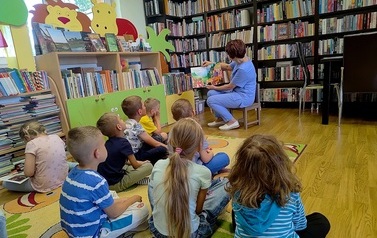 Dzieci siedzą na dywanie i patrzą na Panią Gabrysię, ktora im czyta bajkę obrazkami skierowaną do dzieci