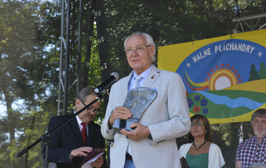 Przed mikrofonem stoi starszy siwy mężczyzna w okularach i trzyma statuetkę Ambasador Kociewia. Za nim z tyłu na scenie stoją inni ludzie.
