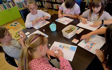 Dzieci siedzą przy stoliku i na białych kartkach wklejają obrazki dotyczące powstawania książki.