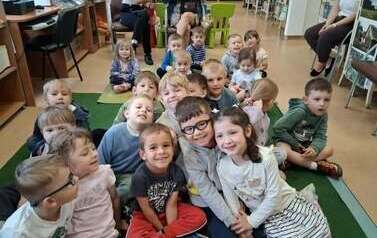 Dzieci siedzą na dywanie i słuchają bajki kt&oacute;rą czyta bibliotekarka