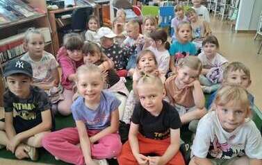Dzieci siedzą na dywanie i słuchają bajki kt&oacute;rą czyta bibliotekarka