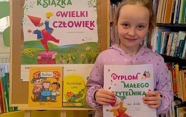 Uśmiechnięta dziewczynka w blond włosach stoi w bibliotece w Pinczynie przy tablicy z plakatem projektu i pokazuje sw&oacute;j nowy dyplom Małego Czytelnika.