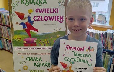 Uśmiechnięty chlopiec w blond włosach stoi w Bibliotece w Zblewie  przy tablicy z plakatami Mała Książka Wielki Człowiek i pokazuje sw&oacute;j dyplom Małego Czytelnika.