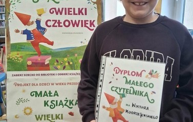 Uśmiechnięty chłopiec w okularach stoi w bibliotece w Zblewie przy tablicy z plakatami Mała Książka Wielki Człowiek i pokazuje sw&oacute;j dyplom