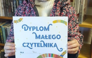 Uśmiechnięta dziewczynka w blond włosach stoi w bibliotece w Borzechowie i pokazuje sw&oacute;j nowy dyplom Małego Czytelnika.