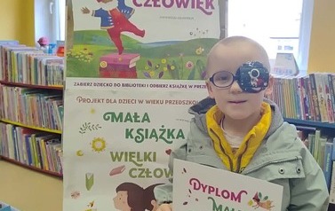 Chłopiec w okularach stoi w bibliotece w Zblewie przy tablicy z plakatami dotyczącemi projektu Mała Książka Wielki Człowiek i trzyma sw&oacute;j nowy dyplom