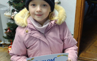 Dziewczynka w r&oacute;żowej kurtce i granatowej czapce pokazuje dyplom w bibliotece w Borzechowie. Za nią stoi sztuczna choinka.