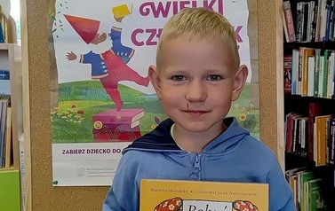 Chlopiec w blond wlosach pokazuje swoją nową książkę pt Bobuś, za nim stoi korkowa tablica z plakatem projektu Mała Książka Wielki Człowiek
