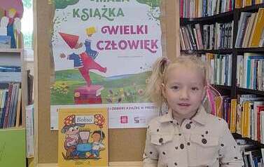 Dziewczynka w blond włosach w kitkach pokazuje swoją nową książkę, za nią stoi korkowa tablica z plakatem projektu Mała Książka Wielki Człowiek