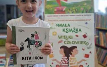Uśmiechnięta dziewczynka w blond wlosach pokazuje swoja nową książkę pt Rita i koń. Za nią stoi tablica z plakatami projektu Mała Książka Wielki Człowiek